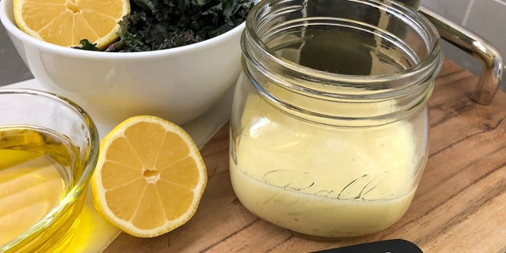 Champagne Garlic Vinaigrette Recipe served on a round mason jar on top of a wooden tray next to fresh lemons oil and a white bowl with leafy greens