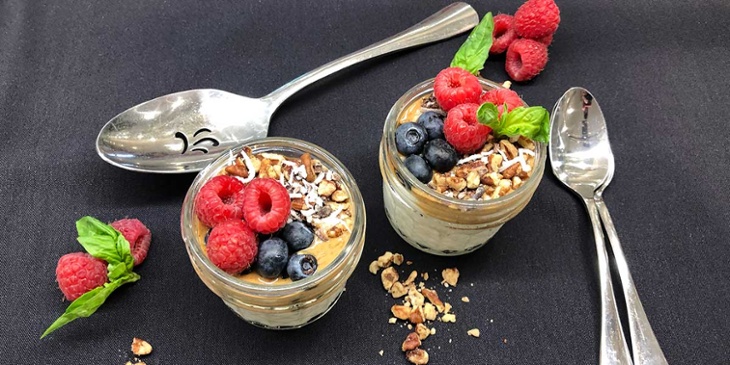 Fully loaded keto breakfast parfait on mason jars surrounded by fresh berries and spoons on a black background