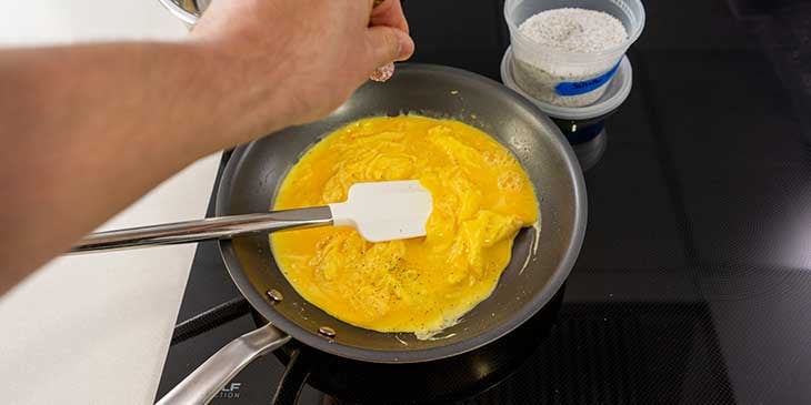 Scrambled eggs being seasoned with salt and pepper as they are cooked on a non-stick pan