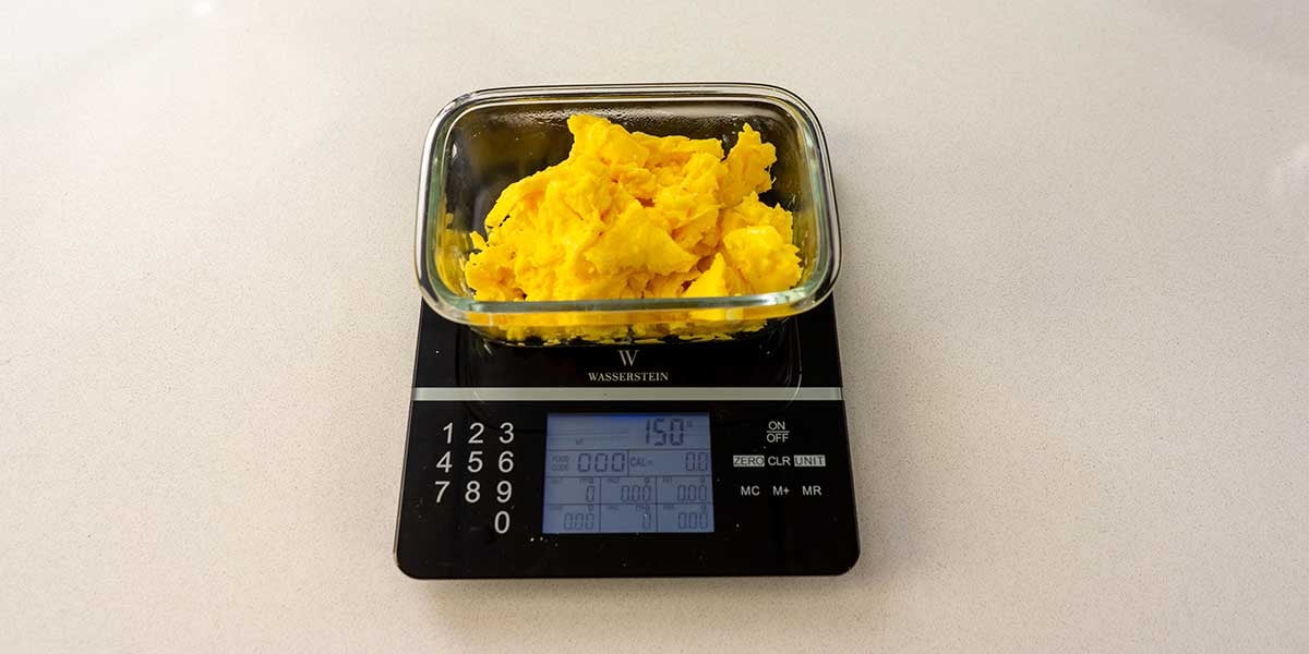 Scrambled eggs portioned into glass rectangular meal prep containers on a black food scale