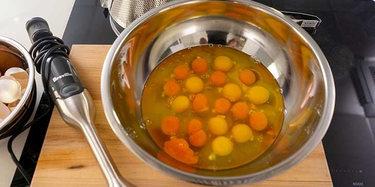 All needed eggs cracked into a stainless steel bowl placed on top of a cutting board next to a Breville hand immersion blender