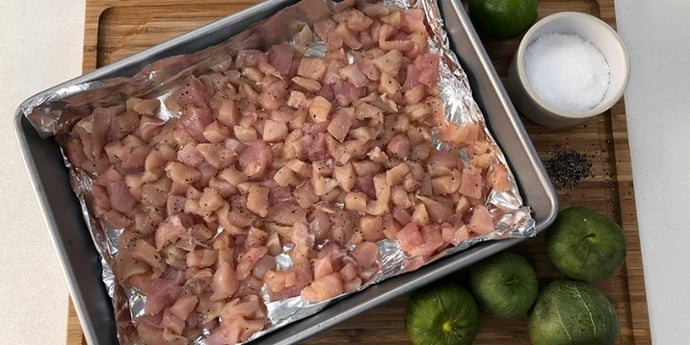 Raw diced chicken on a foil-lined baking sheet placed on top of a wood cutting board