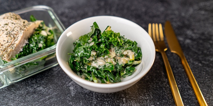Paleo-Aromatic Greens with coconut Recipe plated on a white bowl and meal prep container next to golden utensils on a black backdrop