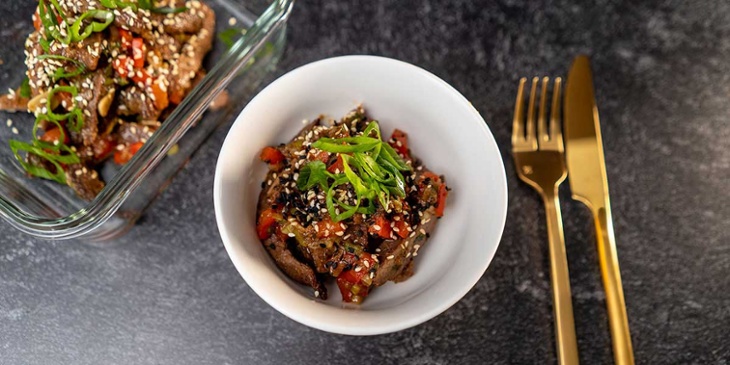 Paleo Mongolian Beef Recipe plated on a small white bowl and on a clear glass meal prep container next to golden utensils on a black backdrop