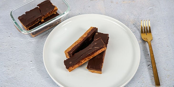 Paleo No-Bake Protein Bars Recipe plated on a white plate and on a glass meal prep container next to a golden fork