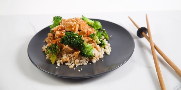 Sriracha Honey Sesame Chicken Trifecta plated on a black small plate with wooden chopsticks