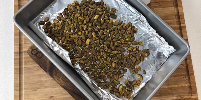 Toasted pistacchio nuts on a foiled-lined baking sheet placed on top of a wooden cutting board