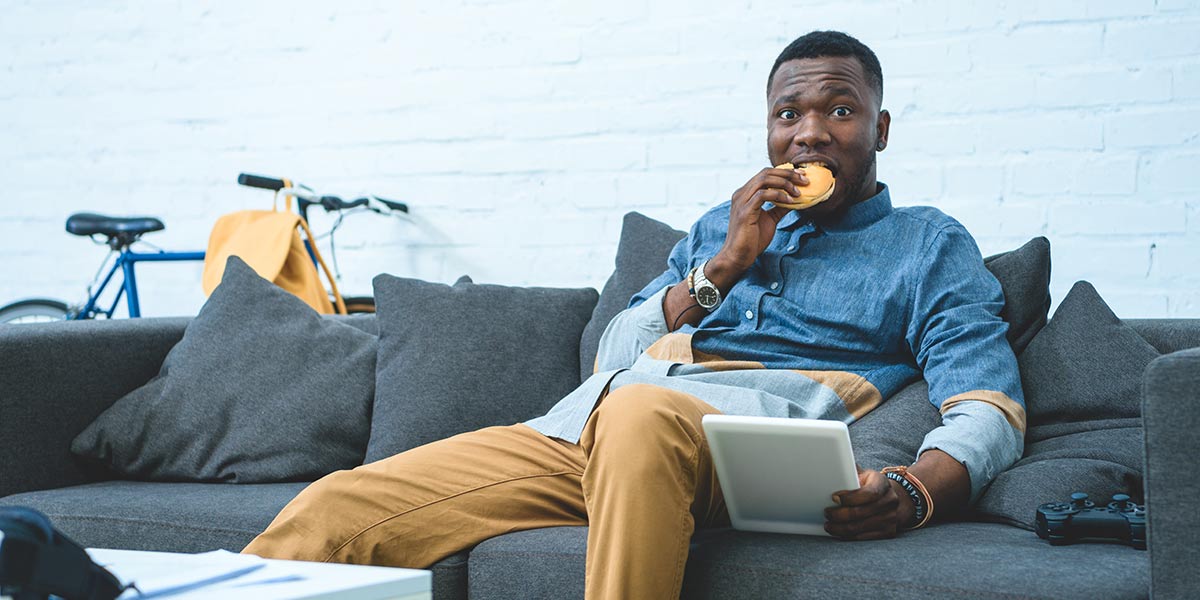 Food's Carbon Footprint: 9 Ways to Reduce Greenhouse Emissions: man eating hamburger with tablet on sofa with bicycle in background
