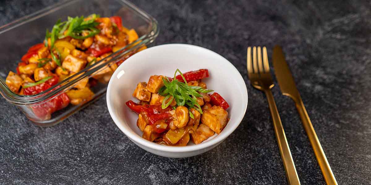 Paleo Cashew Chicken Recipe served on a white bowl and a meal prep container with golden silverware placed on a black background