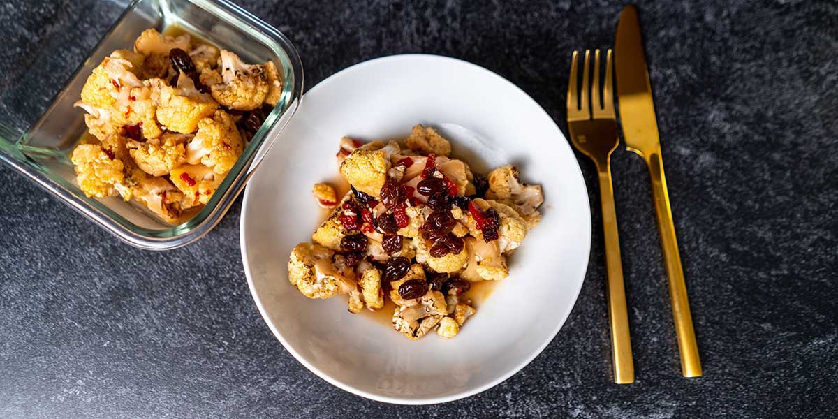 Roasted Paleo Cauliflower with Agrodolce Sauce Recipe served on a white round bowl and a rectangular meal prep container with golden silverware on top of a black background